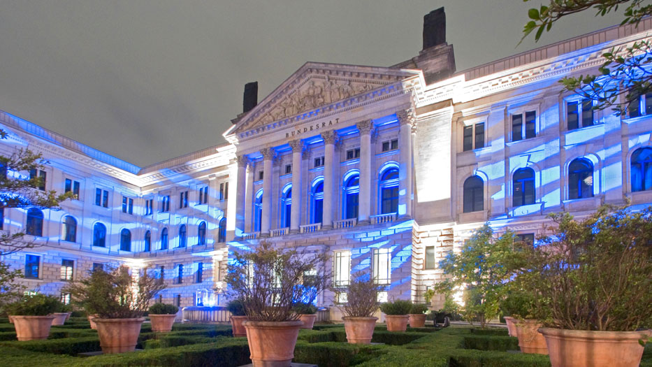Das Bundesratsgebäude ist blau angeleuchtet. Im Vordergrund sind noch einige zu Kugeln geschnittene Büsche im Vorgarten des Gebäudes zusehen.