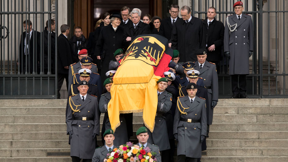 Sargträger - begleitet von der Totenwache - tragen den mit der Bundesdienstflagge zugedeckten Sarg die Treppe am Berliner Dom herunter. Voran gehen zwei Kranzträger mit Blumenschmuck. Im Hintergrund begleitet der damalige Bundespräsident Gauck die Witwe.